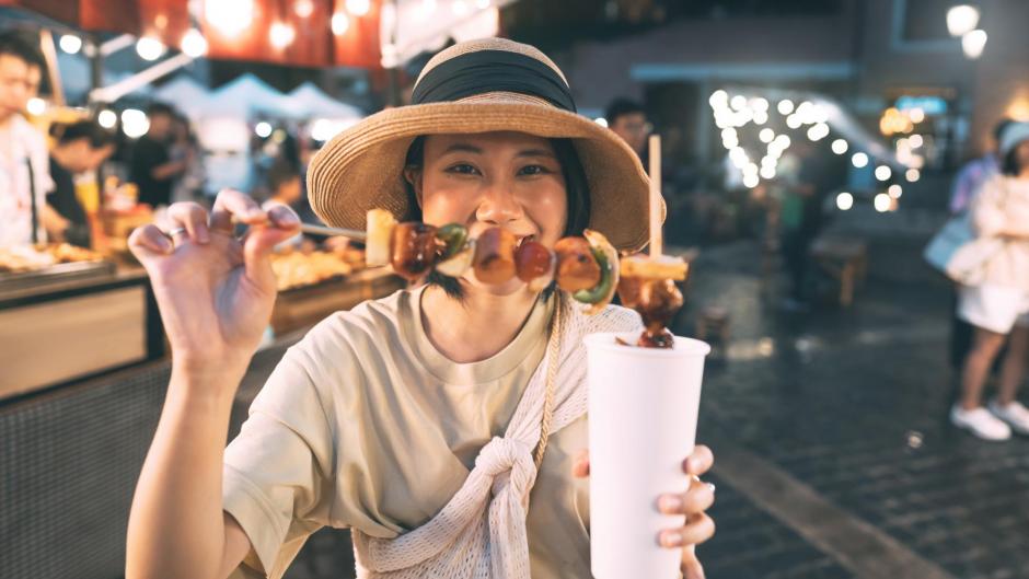 Happy young adult asian foodie woman holding bbq grilled skewers at outdoor vendor night market.