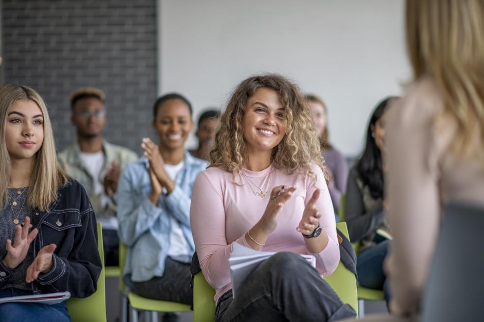 A diverse group of attractive, young, and studious students sits in a room for a lecture from their professor. They are attentive and focused on what's going on. Some are taking notes.